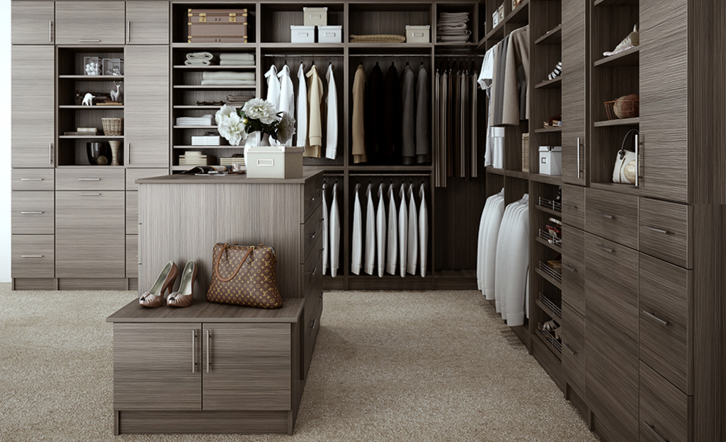 Shoes and a purse sit on an island in the middle of a large gray walk-in closet with wall units of drawers and shelves.