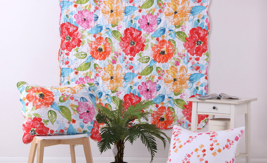 A floral quilt hung as wall art beside a chair with a matching pillow, a small plant, an end table and another pillow.