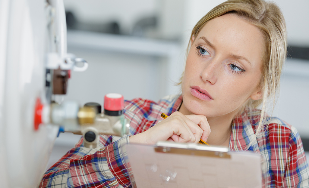 A woman holding a clipboard and a pencil looks closely at a water heater.