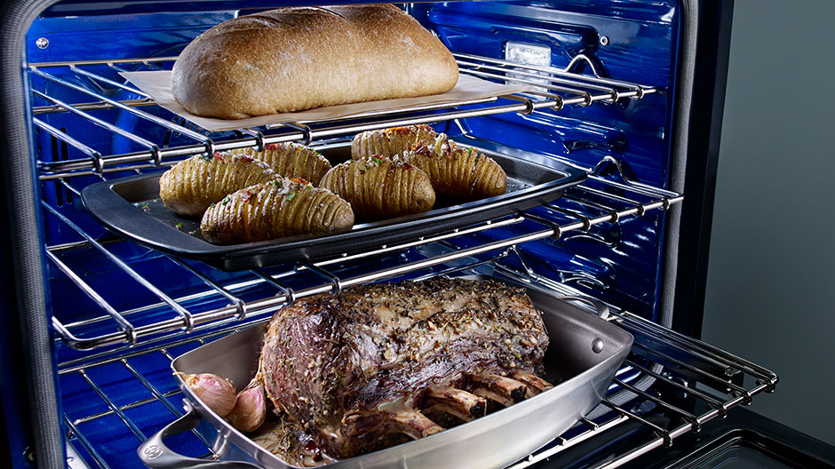 Angled view of open oven showing a large roast on the lower rack, scalloped potatoes on the middle rack and a loaf of bread baking on the upper rack.