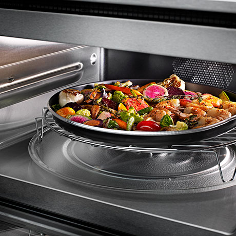 An interior view of the stainless steel convection microwave with a circular rack on the turntable holding a dish of roasting vegetables.
