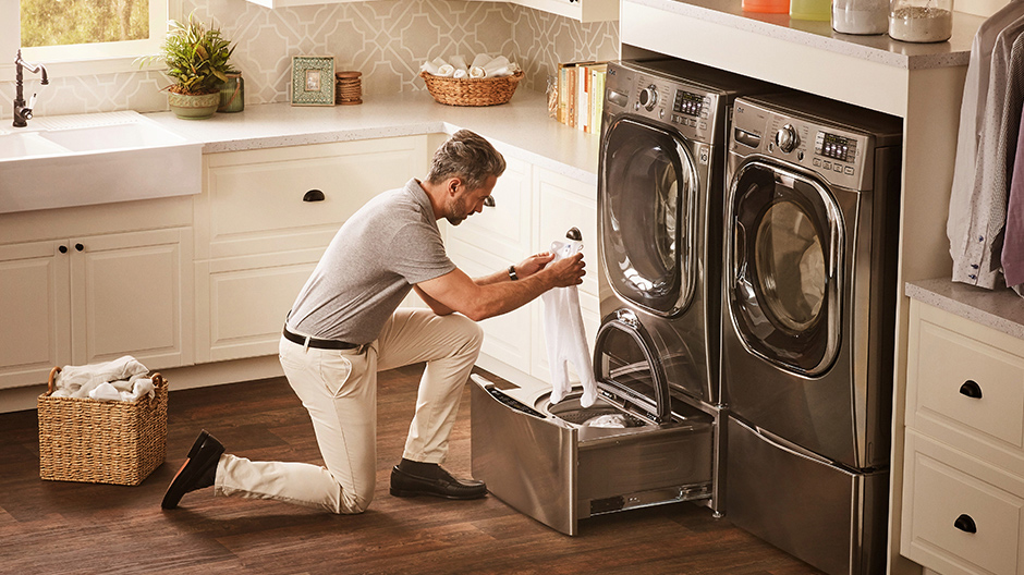 A man kneels in front of LG TWINWash in a bright laundry room, loading a babyÕs onesie into the LG SideKick Pedestal Washer