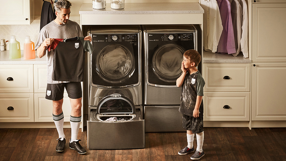 Sporty dad holds a dirty soccer uniform up to his son while they stand in front of the LG TWINWash with the LG SideKick Pedestal Washer