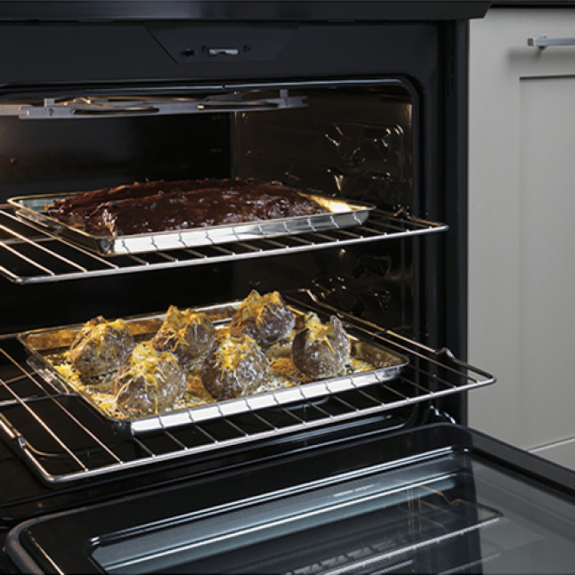 Two metal racks with food baking on them are pulled halfway out of the oven's spacious cavity. The oven door has been lowered.