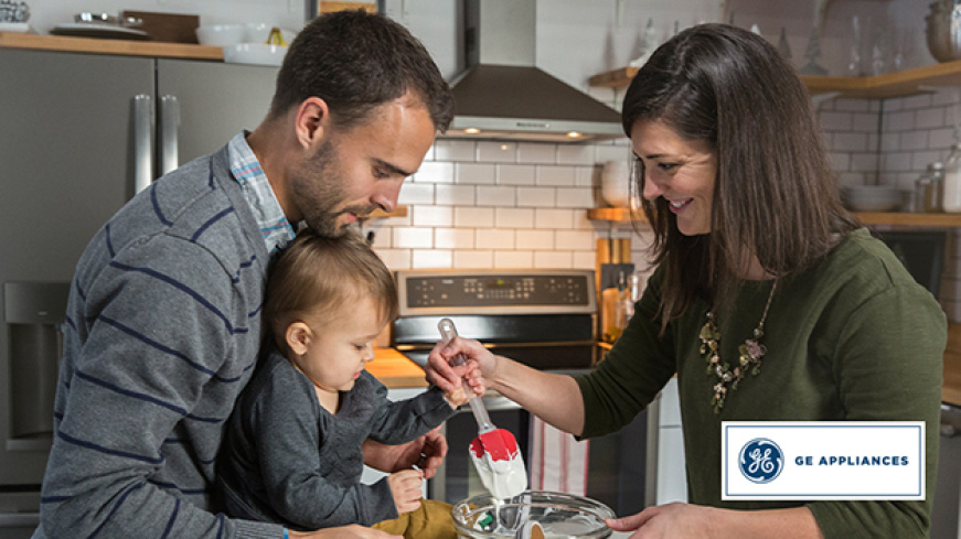 A family is in their kitchen together. The father holds the child up as the smiling mother guides the child's hand to stir a glass bowl of batter. A family is in their kitchen together. The father holds the child up as the smiling mother guides the child's hand to stir a glass bowl of batter.