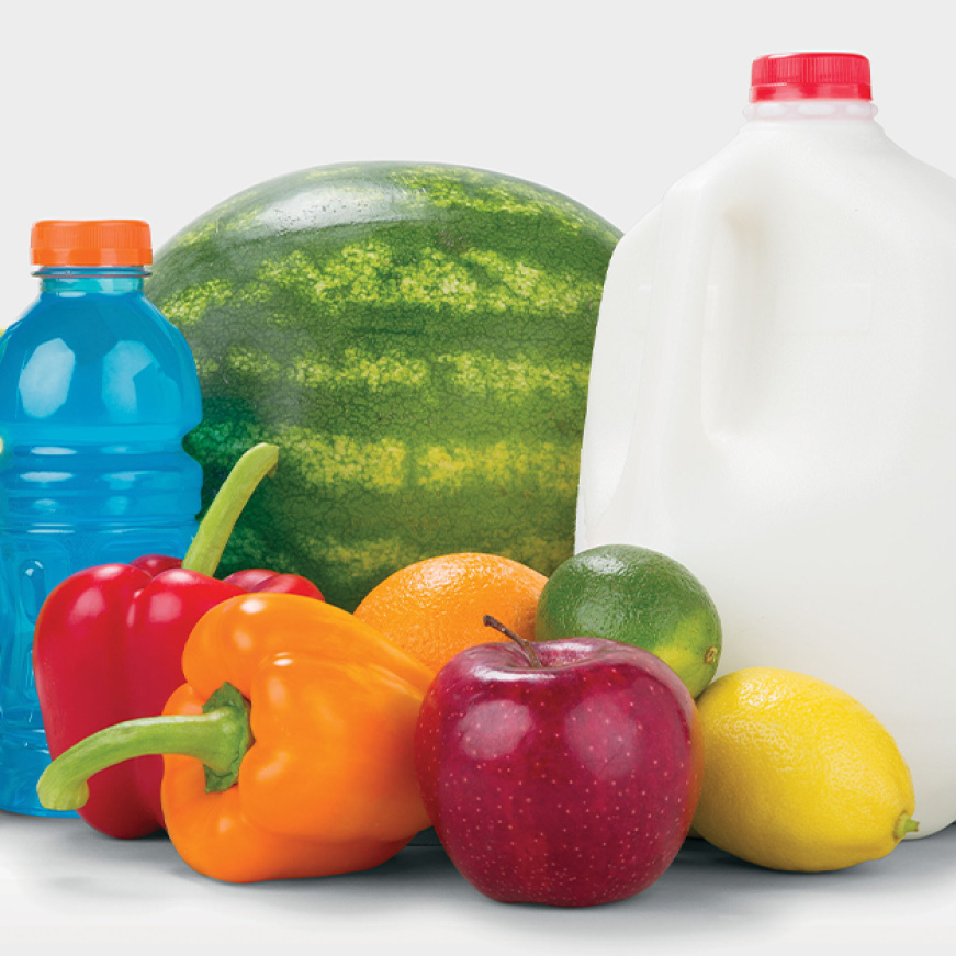 A variety of food sits on a white backdrop. Apples, peppers, and citrus are arranged in front of a sports drink, a gallon of milk, and a watermelon