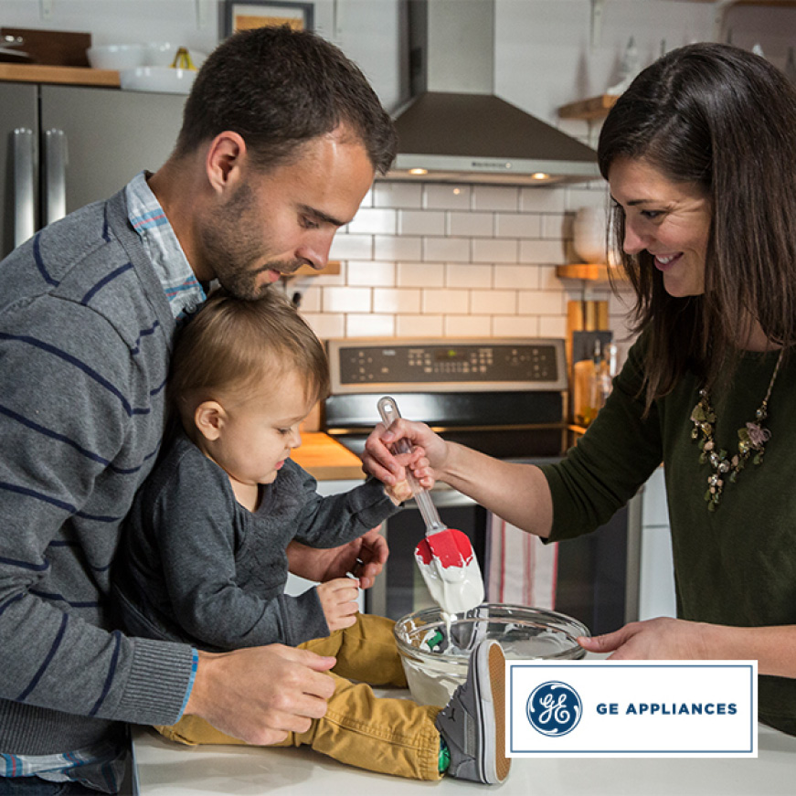 A family is in their kitchen together. The father holds the child up as the smiling mother guides the child’s hand to stir a glass bowl of batter. A family is in their kitchen together. The father holds the child up as the smiling mother guides the child’s hand to stir a glass bowl of batter.