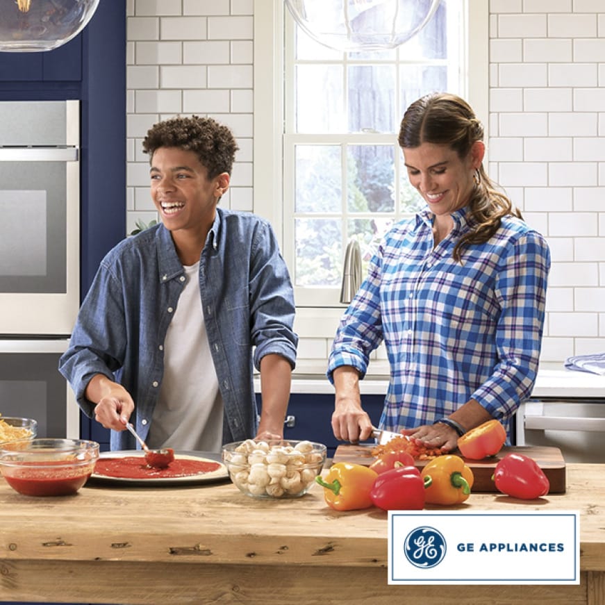A woman chops peppers while a laughing teenage boy spoons pizza sauce on to a pizza crust. A woman chops peppers while a laughing teenage boy spoons pizza sauce on to a pizza crust.