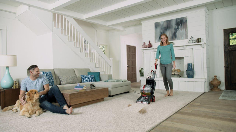 A woman using the Power Scrub Elite Pet Plus Carpet Cleaner in a living room with a man and dog sitting near the couch. A woman using the Power Scrub Elite Pet Plus Carpet Cleaner in a living room with a man and dog sitting near the couch.