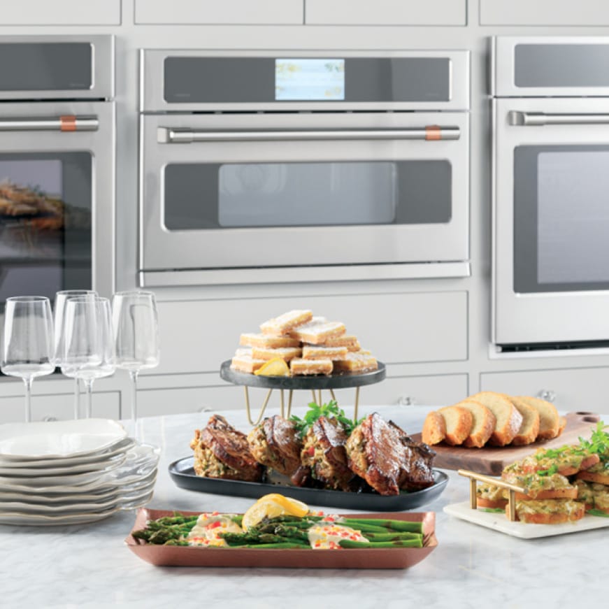 An advantium oven is installed between two wall ovens. A variety of gourmet foods are arranged on a counter in front of the ovens.