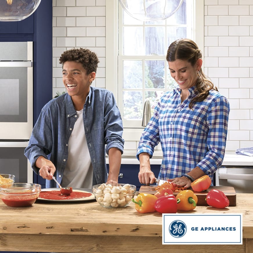 A woman chops peppers while a laughing teenage boy spoons pizza sauce on to a pizza crust. A woman chops peppers while a laughing teenage boy spoons pizza sauce on to a pizza crust.