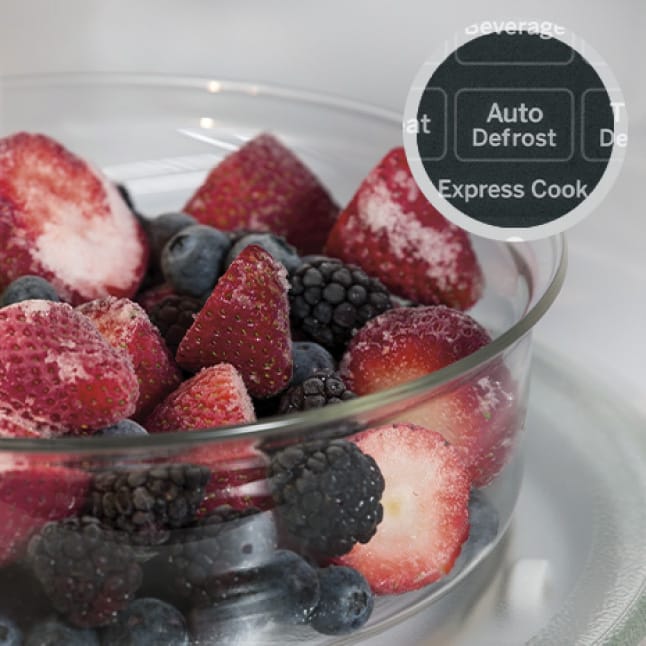 Frozen berries in a glass bowl defrost inside the microwave’s cavity.A circular overlay shows the defrost button on the control panel.